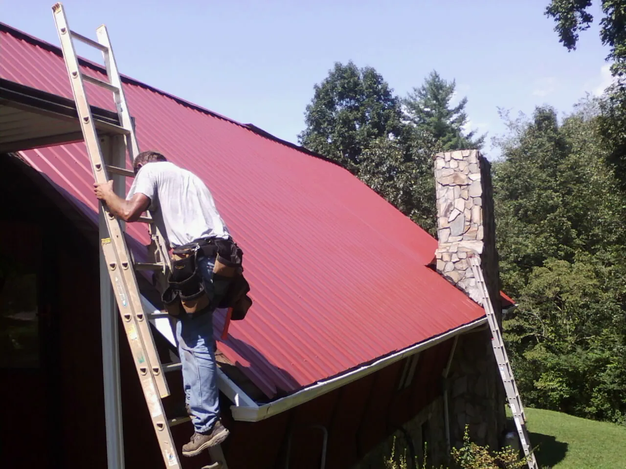 Roofer on a ladder beside a red metal roof and stone chimney — Soffit & Fascia Repair by Roofing Brothers in Osage City